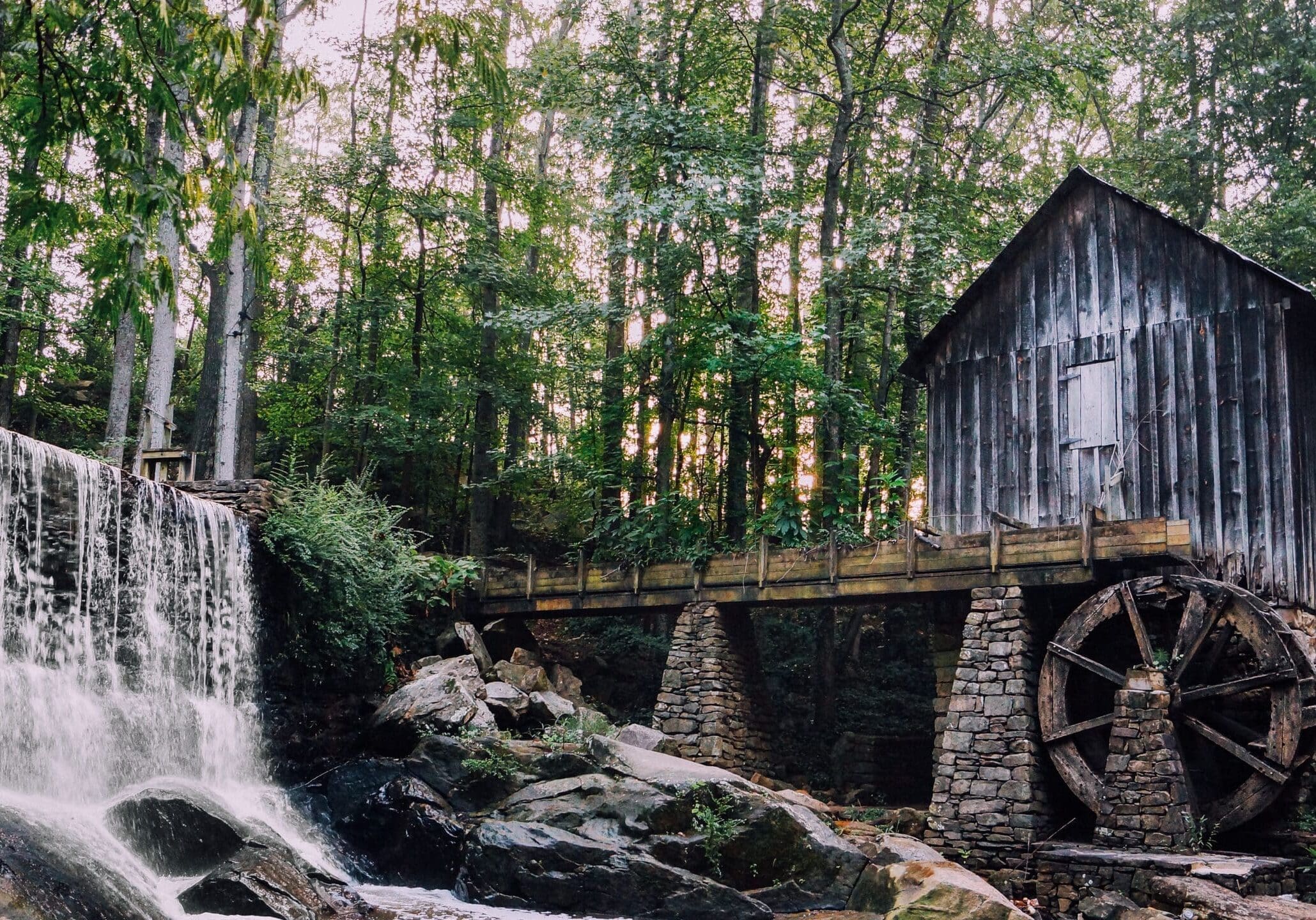 Historic wooden water mill with a large stone foundation and water wheel beside a cascading waterfall in Marietta, Georgia forest.